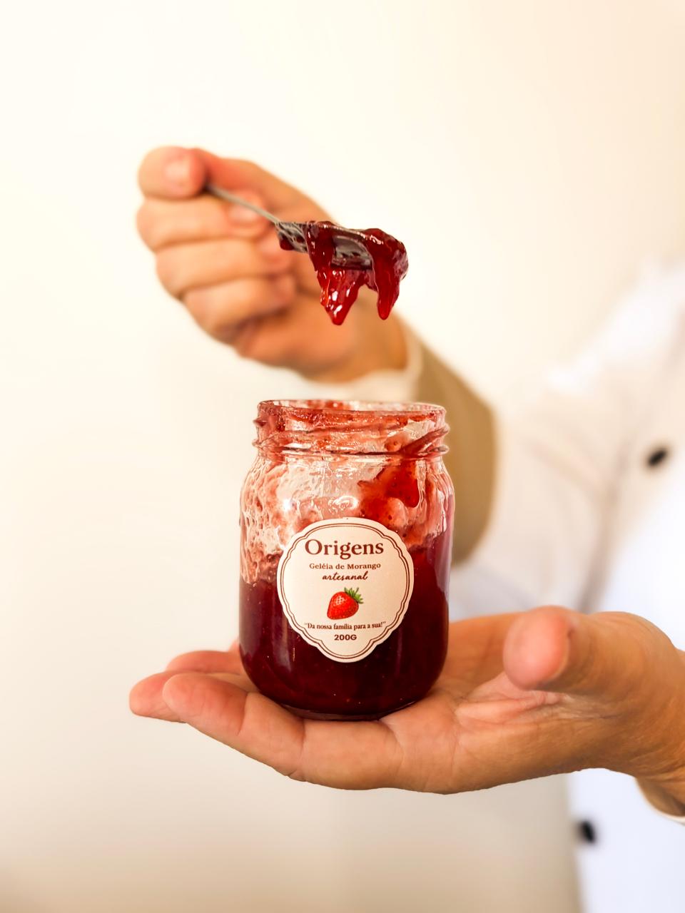 Pote de geleia artesanal de morango Origens na mão, com colher cheia de geleia